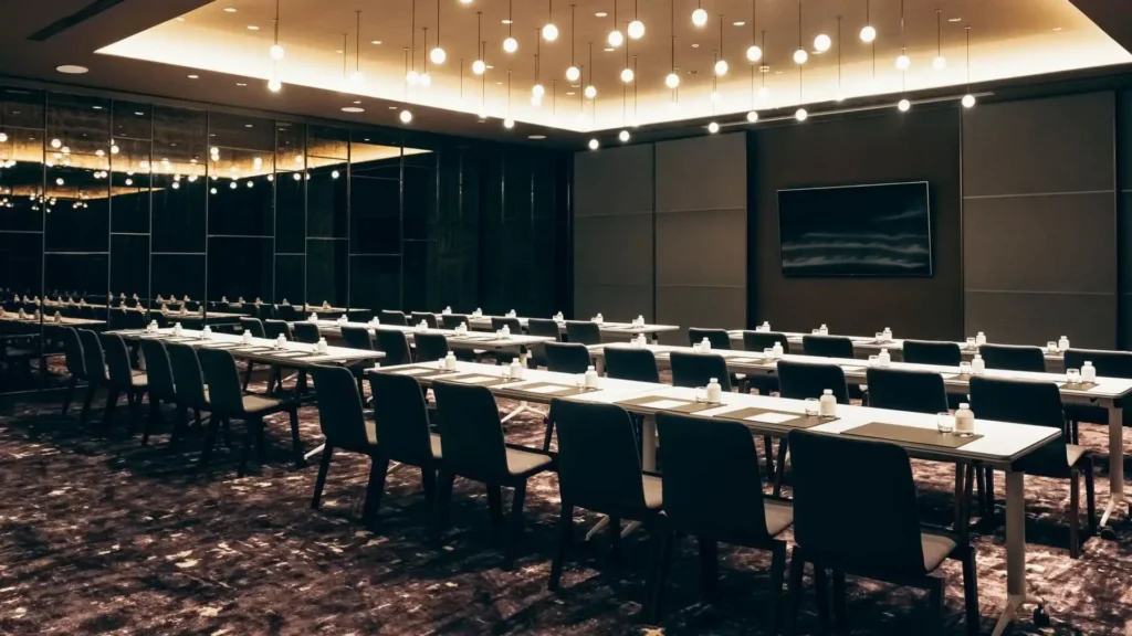 A modern conference room with rows of tables and chairs, set with notepads and bottled water, under ceiling lights.